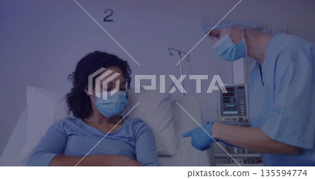 Leaning gloved nurse in blue scrubs pointing at patient arm on hospital bed, with infusion pump 135594774