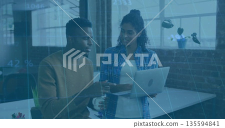 Collaborating coworkers in casual attire discussing project at office desk with laptop and mug 135594841