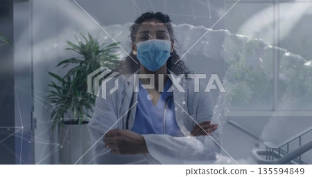 Standing masked doctor crossing arms in corridor with stethoscope, displaying network overlay Standing masked doctor crossing arms in corridor with stethoscope, displaying network overlay 135594849