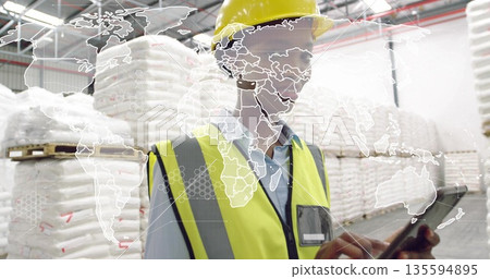 Worker wearing safety gear reviewing inventory on tablet in warehouse, with bulk bags, map overlay 135594895