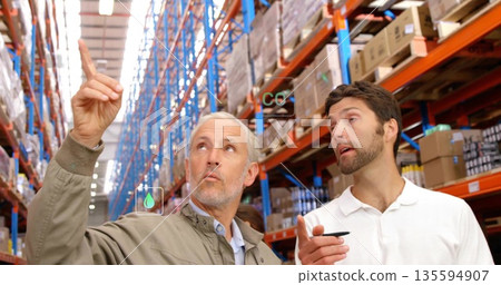 Inspecting two male colleagues pointing toward tall racks inside warehouse, with boxes, copy space 135594907