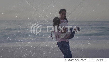 Twirling mother wearing pink plaid shirt holding daughter in white jacket at beach, sparkle overlay 135594919