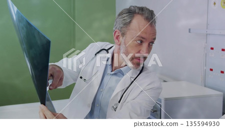 Pointing doctor in lab coat holding radiograph film on wall in exam room, with stethoscope, cabinet Pointing doctor in lab coat holding radiograph film on wall in exam room, with stethoscope, cabinet 135594930