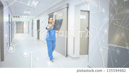 Nurse in blue scrubs holding and inspecting X-ray film in hospital corridor, with network overlay 135594982