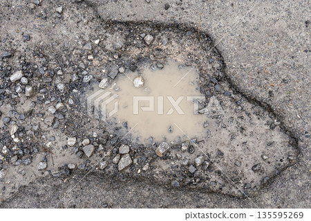 Muddy puddle filling a pothole on a damaged asphalt road surrounded by cracked pavement and loose gravel. Concept of urban infrastructure deterioration 135595269