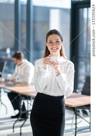 Cute young business woman enjoying her tea and looking contented Cute young business woman enjoying her tea and looking contented 135595624
