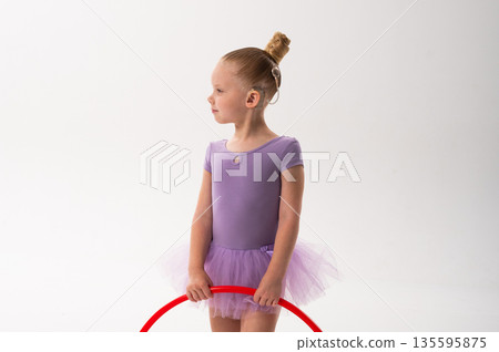 A smiling ballerina with a cochlear implant raises a red hoop in a ballet pose. Copy space. Inclusive sport, hearing technology, and confident childhood expression through rhythmic dance and movement. A smiling ballerina with a cochlear implant raises a red hoop in a ballet pose. Copy space. Inclusive sport, hearing technology, and confident childhood expression through rhythmic dance and movement. 135595875