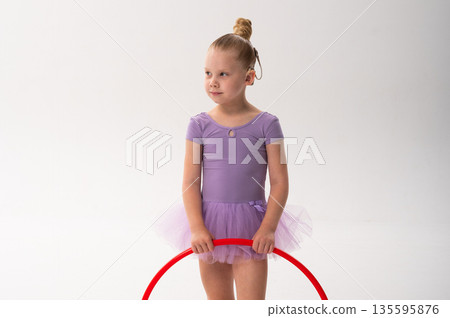 A smiling ballerina with a cochlear implant raises a red hoop in a ballet pose. Inclusive sport, hearing technology, and confident childhood expression through rhythmic dance and movement. 135595876