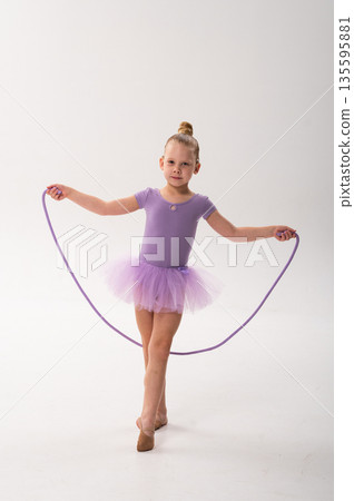 Young ballerina with a cochlear implant stretches a red hoop while posing in a lavender tutu. Inclusive gymnastics, hearing technology, and childhood coordination through rhythmic dance and movement. Young ballerina with a cochlear implant stretches a red hoop while posing in a lavender tutu. Inclusive gymnastics, hearing technology, and childhood coordination through rhythmic dance and movement. 135595881