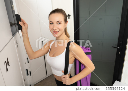 Cute blonde young woman with a violet bag opening locker in gym 135596124