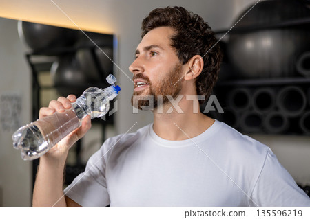 Dark-haired caucasian young man in the gym with a bottle of water in hands 135596219