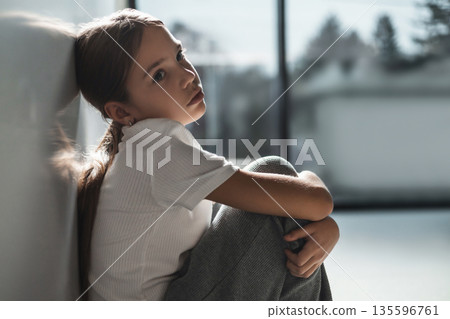 Child sitting by window gazing outward looks sad and unhappy 135596761