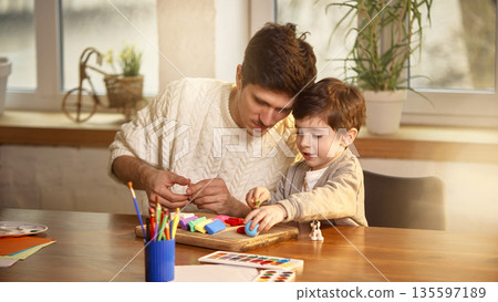 Father and young son concentrating on clay modeling at wooden table. Father and young son concentrating on clay modeling at wooden table. 135597189