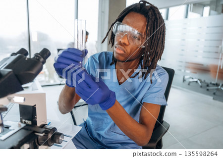 In protective eyewear, with microscope. Black man doctor is in the office In protective eyewear, with microscope. Black man doctor is in the office 135598264