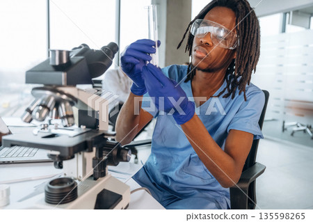In protective eyewear, with microscope. Black man doctor is in the office In protective eyewear, with microscope. Black man doctor is in the office 135598265