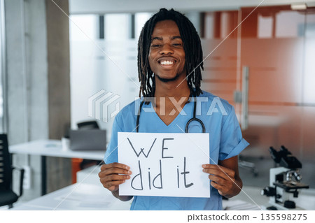 Holding paper with text 'we did it'. Black man doctor is in the office 135598275