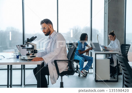 In lab coat, sitting with microscope. Doctors are in the clinic In lab coat, sitting with microscope. Doctors are in the clinic 135598420
