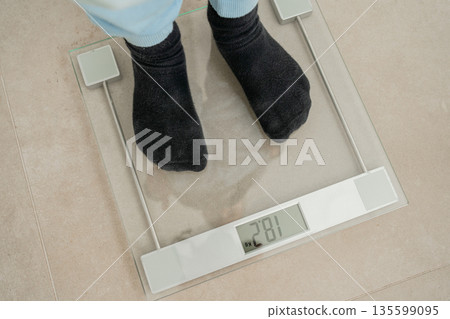 Person standing on digital scale at home interior. Concept of healthy habits, weight control and wellness lifestyle Person standing on digital scale at home interior. Concept of healthy habits, weight control and wellness lifestyle 135599095