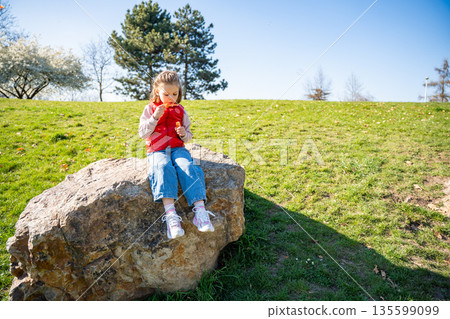 Little girl sitting on decorative stones blowing soap bubbles in city park. Spring childhood leisure and joyful outdoor play. Little girl sitting on decorative stones blowing soap bubbles in city park. Spring childhood leisure and joyful outdoor play. 135599099