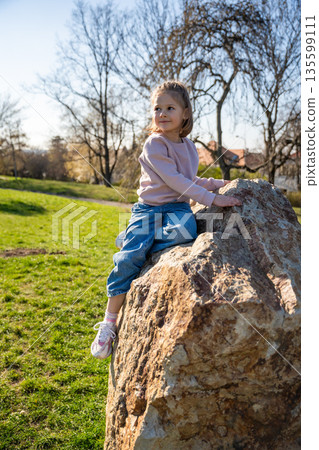 Little girl jumping on decorative stones in city park. Active spring play and outdoor childhood lifestyle. 135599111