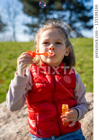 Little girl sitting on decorative stones blowing soap bubbles in city park. Spring childhood leisure and joyful outdoor play. Little girl sitting on decorative stones blowing soap bubbles in city park. Spring childhood leisure and joyful outdoor play. 135599166