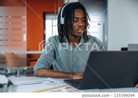 In headphones, by laptop. Black businessman is working in office In headphones, by laptop. Black businessman is working in office 135599394