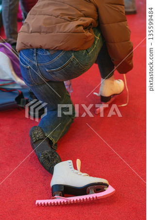 Woman lacing figure skates closeup. Person tying ice skates on red carpet before skating session. Preparing for winter skating on an outdoor ice rink. Winter sport, ice skating activity 135599484