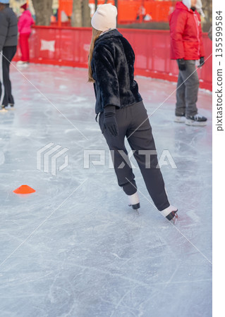 Rear view of woman on ice in black jacket. Woman skating on outdoor ice rink in winter clothes. Concept of sport, winter fun and seasonal recreation Rear view of woman on ice in black jacket. Woman skating on outdoor ice rink in winter clothes. Concept of sport, winter fun and seasonal recreation 135599584