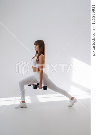 Holding dumbbells and doing squats. Young sportive woman is in the studio 135599931
