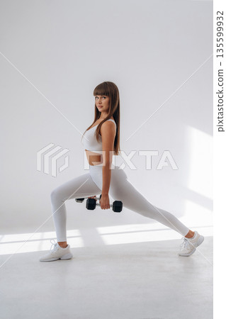 Holding dumbbells and doing squats. Young sportive woman is in the studio 135599932