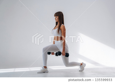 Holding dumbbells and doing squats. Young sportive woman is in the studio 135599933