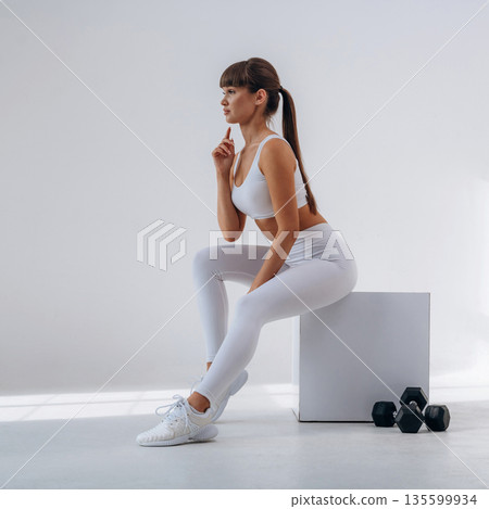 Sitting on the white cube, dumbbells are on the floor. Young sportive woman is in the studio 135599934