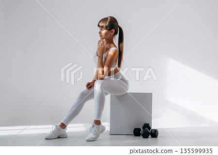 Sitting on the white cube, dumbbells are on the floor. Young sportive woman is in the studio 135599935