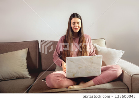 Young woman working on laptop at home while sitting on couch in comfortable clothing shows remote work lifestyle in modern living space with soft lighting Young woman working on laptop at home while sitting on couch in comfortable clothing shows remote work lifestyle in modern living space with soft lighting 135600045