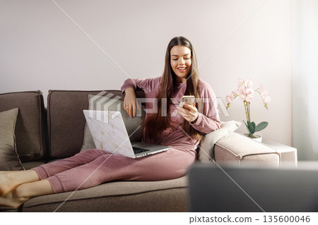 Young woman sits on sofa using smartphone while laptop rests on cushion in cozy room during day Young woman sits on sofa using smartphone while laptop rests on cushion in cozy room during day 135600046