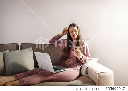 Woman sits on couch with laptop and phone, enjoying a moment of leisure at home in a comfortable setting, absorbing the online world while relaxing at ease Woman sits on couch with laptop and phone, enjoying a moment of leisure at home in a comfortable setting, absorbing the online world while relaxing at ease 135600047