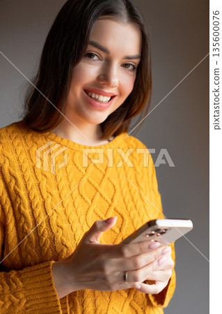 Woman wearing a yellow sweater smiles while holding a smartphone in a softly lit indoor space during the daytime Woman wearing a yellow sweater smiles while holding a smartphone in a softly lit indoor space during the daytime 135600076