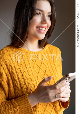 Woman in a yellow sweater smiles while looking at a smartphone in a warm indoor space during the afternoon Woman in a yellow sweater smiles while looking at a smartphone in a warm indoor space during the afternoon 135600077