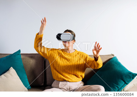 A woman is using a virtual reality headset while moving her hands to engage with the visual content. She is inside a room with light walls and a simple background. A woman is using a virtual reality headset while moving her hands to engage with the visual content. She is inside a room with light walls and a simple background. 135600084