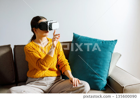 Woman sitting on a couch wearing virtual reality headset while interacting with immersive technology in a modern living room setting during daytime Woman sitting on a couch wearing virtual reality headset while interacting with immersive technology in a modern living room setting during daytime 135600090