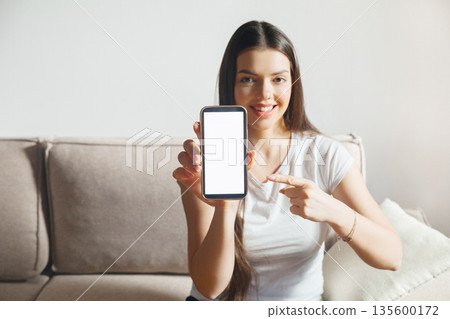 Young woman shows smartphone screen while sitting on a couch in a bright room during the day engaging in a casual activity with a friendly expression Young woman shows smartphone screen while sitting on a couch in a bright room during the day engaging in a casual activity with a friendly expression 135600172