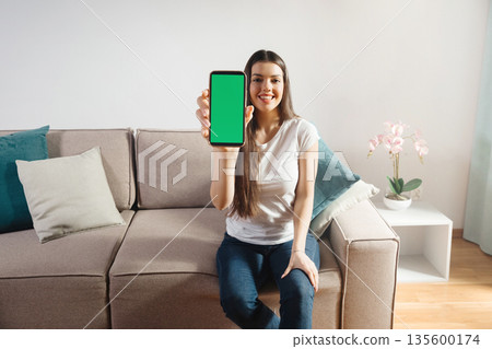 Woman sits on a couch and holds a smartphone displaying a blank green screen in a modern living room environment with decorative plants and cushions 135600174
