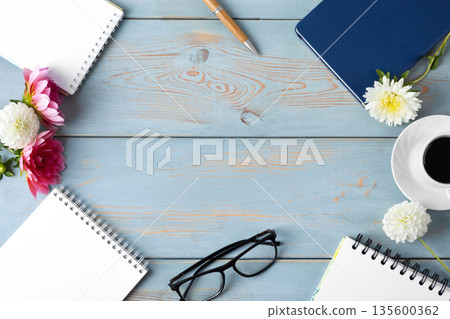 A blue wooden desk with blank notebooks, glasses, a pen, dahlias, and coffee, with a center space available. Morning coffee cup. A blue wooden desk with blank notebooks, glasses, a pen, dahlias, and coffee, with a center space available. Morning coffee cup. 135600362
