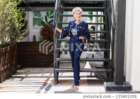 Stylish happy smiling woman stands outdoors on city sidewalk wearing fashionable denim jacket and cigarette jeans. Elegant mature model, modern fashion and urban lifestyle Stylish happy smiling woman stands outdoors on city sidewalk wearing fashionable denim jacket and cigarette jeans. Elegant mature model, modern fashion and urban lifestyle 135601208