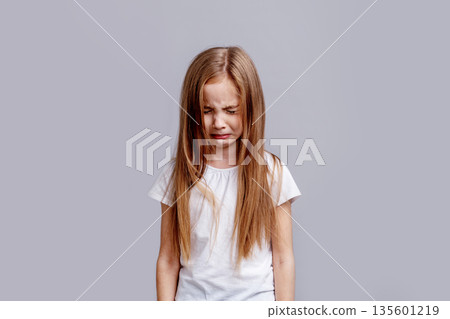 Crying young girl with long blond hair in white T-shirt, eyes closed and face scrunched in tears, isolated on grey background in emotional portrait, sadness and vulnerability 135601219
