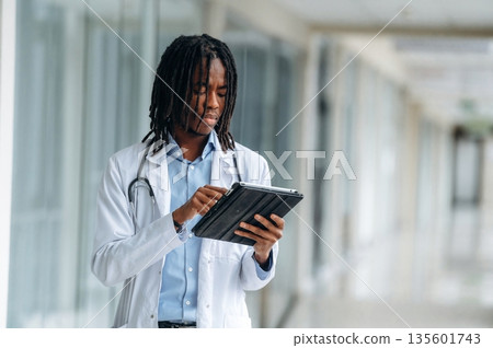 Digital notepad is in hands. Young male black doctor is in the corridor 135601743