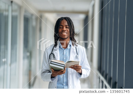 Lot of learning material, holding books. Young male black doctor is in the corridor Lot of learning material, holding books. Young male black doctor is in the corridor 135601745