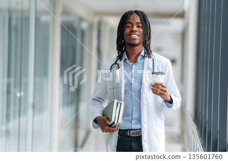 Cup of fresh drink in hand. Young male black doctor is in the corridor 135601760