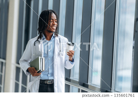 Cup of fresh drink in hand. Young male black doctor is in the corridor 135601761