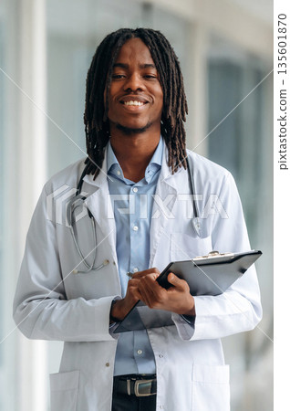 Joyful, positive facial expression. Young male black doctor is in the corridor Joyful, positive facial expression. Young male black doctor is in the corridor 135601870